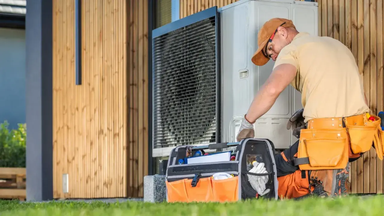 Instalator w pasie narzędziowym sprawdza jednostkę zewnętrzną pompy ciepła przy drewnianym domu.