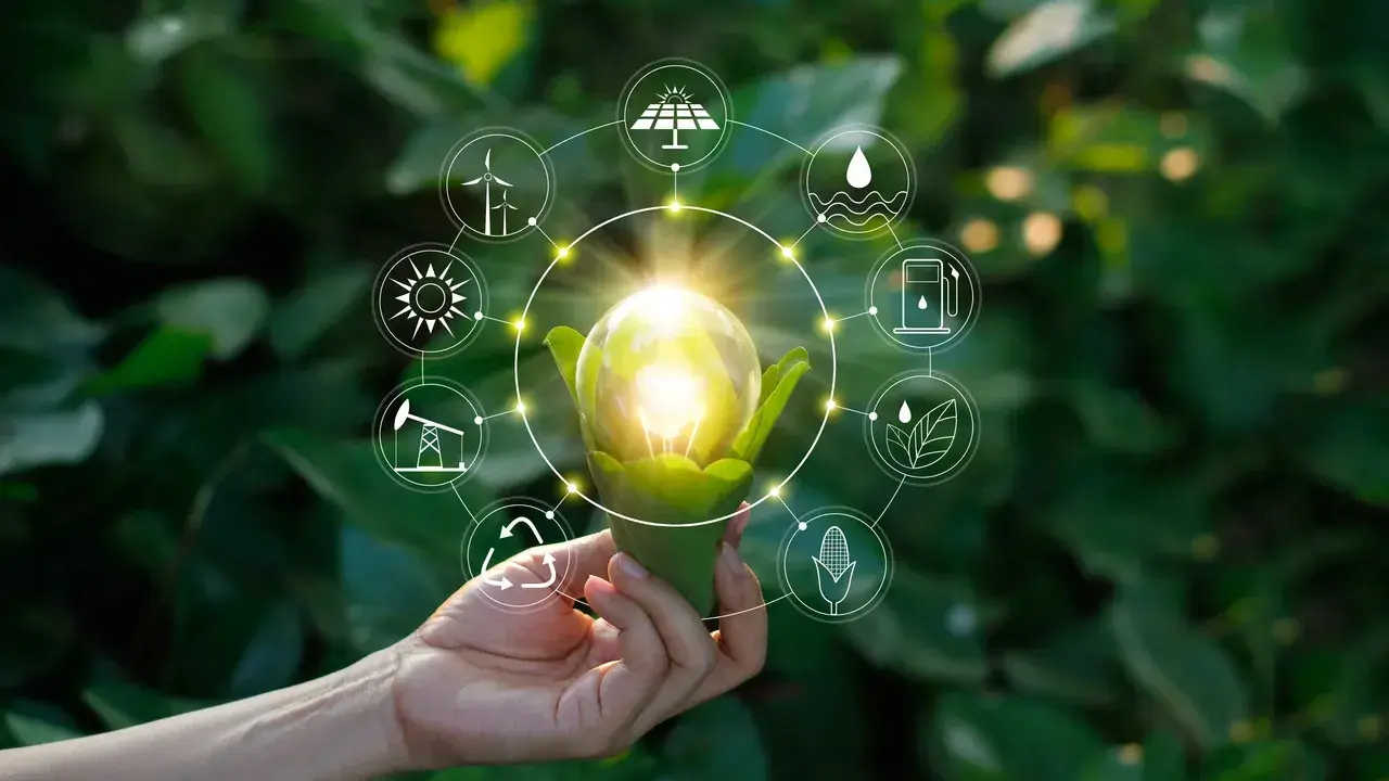 Dłoń trzyma żarówkę z zielonymi liśćmi, otoczoną ikonami symbolizującymi niekonwencjonalne źródła energii: wiatr, słońce, wodę, biomasę i paliwa.