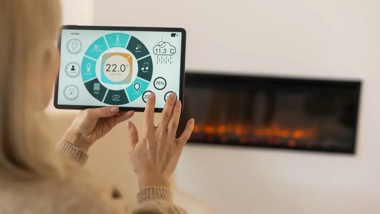 Kobieta steruje inteligentnym domem przez tablet, kontrolując temperaturę i inne funkcje. Widoczny jest ekran z ikonami i wskaźnikami, a w tle kominek.