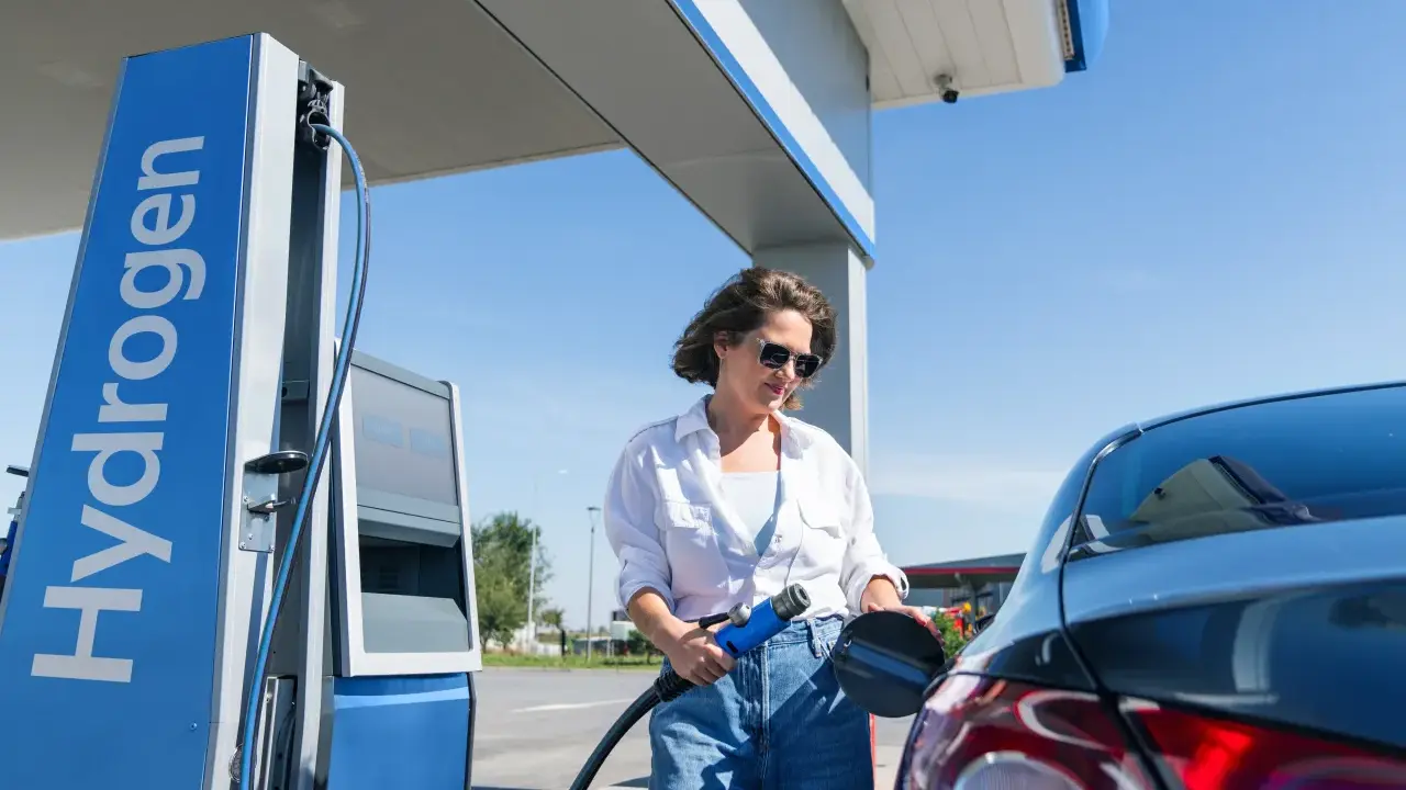 Kobieta tankuje auta na wodór. Stacja z napisem "Hydrogen" i niebieska dystrybutor.