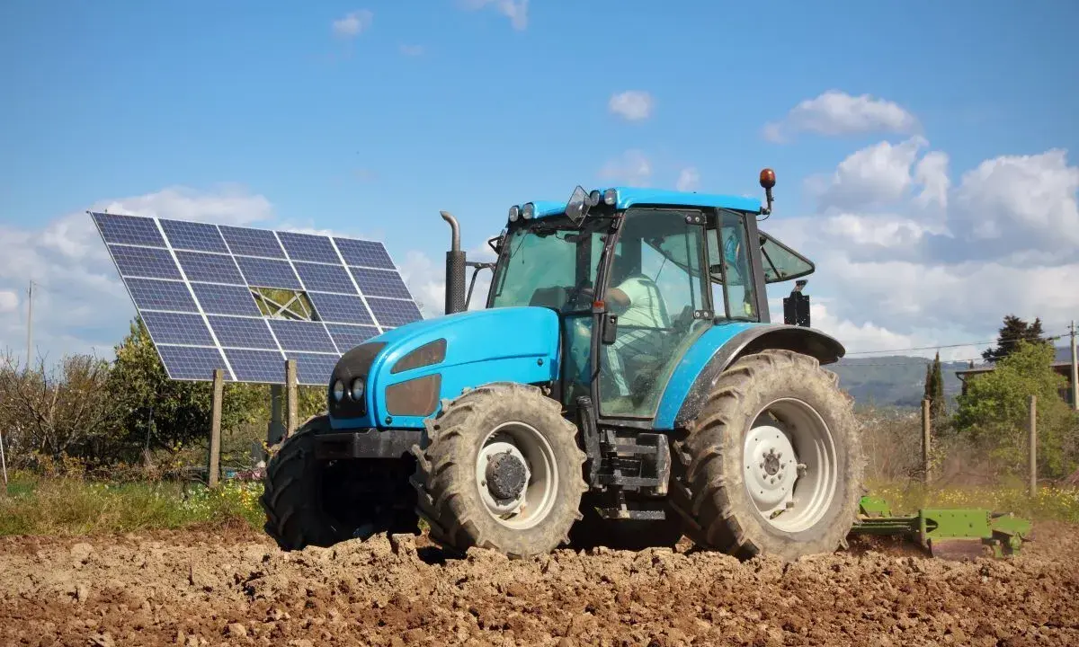 Błękitny traktor pracuje na polu, obok paneli fotowoltaika arimr. Słońce i chmury na niebie.