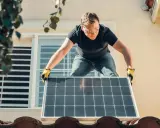 Montaż paneli fotowoltaicznych, czyli mikroinstalacji OZE, na dachu domu. Pracownik w okularach ochronnych i rękawicach ostrożnie układa panel.