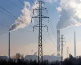Linie energetyczne i dymiące kominy elektrowni na tle błękitnego nieba. W tle widać kilka wież transmisyjnych i budynki przemysłowe.
