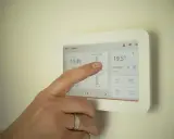 Dłoń dotyka ekranu termostatu, pokazując temperaturę 19,5°C. Ustawienia ogrzewania pomogą określić, jak często powinien włączyć się piec gazowy.