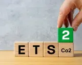 Dłoń układa kostkę z cyfrą 2 na kostce z napisem "CO2", obok kostek z napisami "ETS". Sugeruje to nowy podatek od ogrzewania gazem.