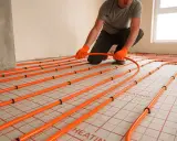 Instalator w pomarańczowych rękawicach układa pomarańczowe rury ogrzewania podłogowego.