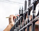 Ręka maluje pędzlem metalową bramę, pokazując zastosowanie farby chlorokauczukowej do ochrony przed rdzą.