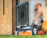 Instalator w pasie narzędziowym sprawdza jednostkę zewnętrzną pompy ciepła przy drewnianym domu.
