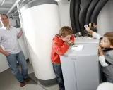 Ojciec z dziećmi przy pompie ciepła. Czy pompę ciepła można wyłączyć? Chłopcy badają urządzenie, ojciec obserwuje.