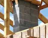 Budowa komina systemowego z bloczków betonowych, gotowego do podłączenia pieca. Drewniana konstrukcja dachu w tle.