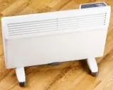 Biały grzejnik elektryczny na kółkach, jeden z rodzajów grzejników elektrycznych, stoi na drewnianej podłodze.