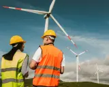 Dwóch pracowników, technik OZE, w kaskach i kamizelkach odblaskowych, obserwuje farmę wiatrową.