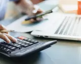 Osoba liczy dzienne zużycie prądu w domu, używając kalkulatora i telefonu. Obok leży laptop i kubek.