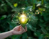 Dłoń trzyma żarówkę z zielonymi liśćmi, otoczoną ikonami symbolizującymi niekonwencjonalne źródła energii: wiatr, słońce, wodę, biomasę i paliwa.