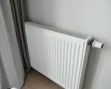 Biały grzejnik panelowy z termostatem, idealny do pokoju 16m2. Obok zasłony.