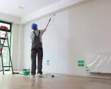 Malowanie ścian wałkiem. Pracownik w kombinezonie i kasku maluje białą ścianę. Obok drabina i wiadro z farbą. Sprawdź, ile za m2 malowania.