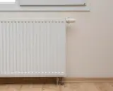 Biały grzejnik przy oknie, termostat ustawiony na minimum. Piec nie podaje ciepła na grzejniki, co oznacza zimne mieszkanie.