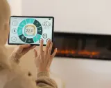 Kobieta steruje inteligentnym domem przez tablet, kontrolując temperaturę i inne funkcje. Widoczny jest ekran z ikonami i wskaźnikami, a w tle kominek.