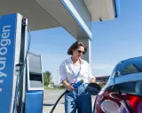 Kobieta tankuje auta na wodór. Stacja z napisem "Hydrogen" i niebieska dystrybutor.