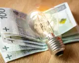 Żarówka na stosie banknotów 100 zł symbolizuje wysokie koszty energii i potrzebę dopłat do prądu.