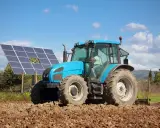 Błękitny traktor pracuje na polu, obok paneli fotowoltaika arimr. Słońce i chmury na niebie.