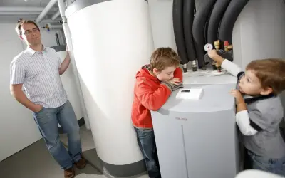 Ojciec z dziećmi przy pompie ciepła. Czy pompę ciepła można wyłączyć? Chłopcy badają urządzenie, ojciec obserwuje.