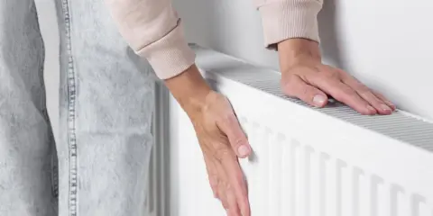 Dłonie sprawdzają temperaturę grzejnika, który słabo grzeje. Zimno w domu, trzeba coś z tym zrobić.