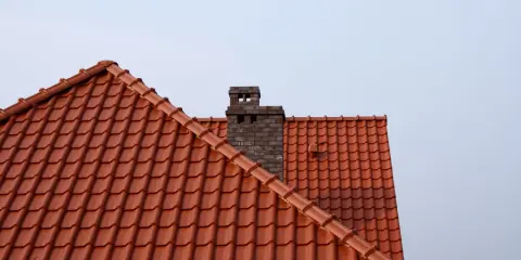 Ceglany komin wystaje ponad dach z czerwonej dachówki. Wlot do komina z pieca znajduje się na wysokości dachu.