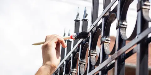 Ręka maluje pędzlem metalową bramę, pokazując zastosowanie farby chlorokauczukowej do ochrony przed rdzą.