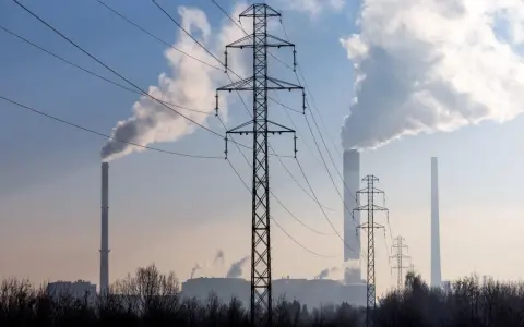 Linie energetyczne i dymiące kominy elektrowni na tle błękitnego nieba. W tle widać kilka wież transmisyjnych i budynki przemysłowe.