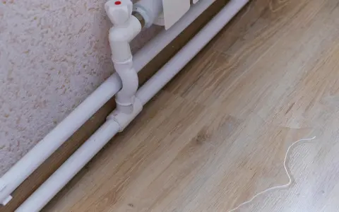 Białe rury do grzejników prowadzą do zaworu. Na drewnianej podłodze widać kałużę wody, co może sugerować problem z instalacją.