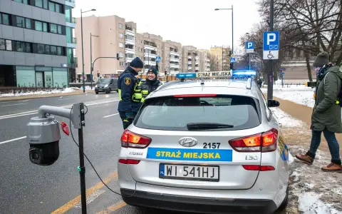 Strażnicy miejscy w Warszawie przy radiowozie i kamerze monitoringu. Dbają o czystą strefę i porządek.