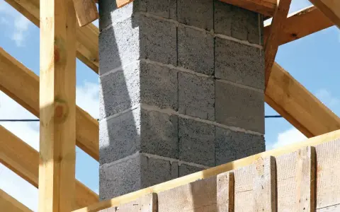 Budowa komina systemowego z bloczków betonowych, gotowego do podłączenia pieca. Drewniana konstrukcja dachu w tle.