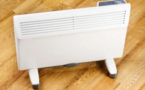 Biały grzejnik elektryczny na kółkach, jeden z rodzajów grzejników elektrycznych, stoi na drewnianej podłodze.
