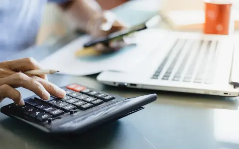 Osoba liczy dzienne zużycie prądu w domu, używając kalkulatora i telefonu. Obok leży laptop i kubek.