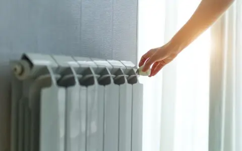 Dłoń reguluje termostat na grzejniku, pokazując różne rodzaje kaloryferów i kontrolę temperatury w domu.