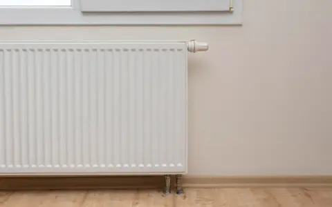 Biały grzejnik przy oknie, termostat ustawiony na minimum. Piec nie podaje ciepła na grzejniki, co oznacza zimne mieszkanie.