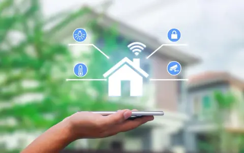 Dłoń trzyma smartfon, z którego wyłania się symbol domu z ikonami oświetlenia, temperatury, zamka i kamery. To wizualizacja systemu zarządzania energią.