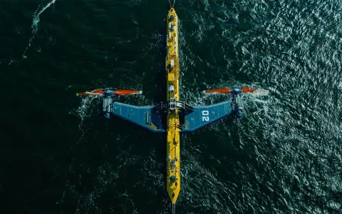 Podwodna turbina wykorzystująca energię pływów, z charakterystycznymi żółto-niebieskimi elementami i pomarańczowymi śmigłami, zanurzona w ciemnych wodach oceanu.