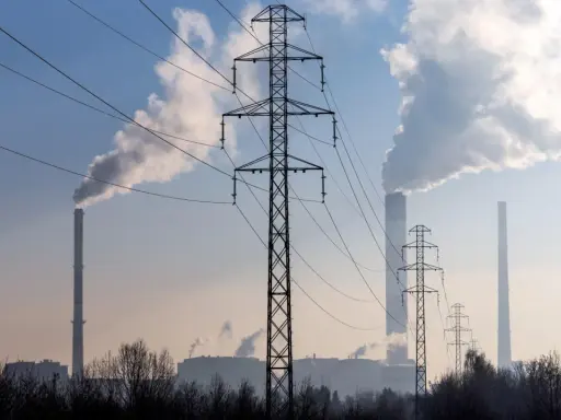 Linie energetyczne i dymiące kominy elektrowni na tle błękitnego nieba. W tle widać kilka wież transmisyjnych i budynki przemysłowe.
