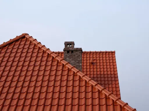 Ceglany komin wystaje ponad dach z czerwonej dachówki. Wlot do komina z pieca znajduje się na wysokości dachu.