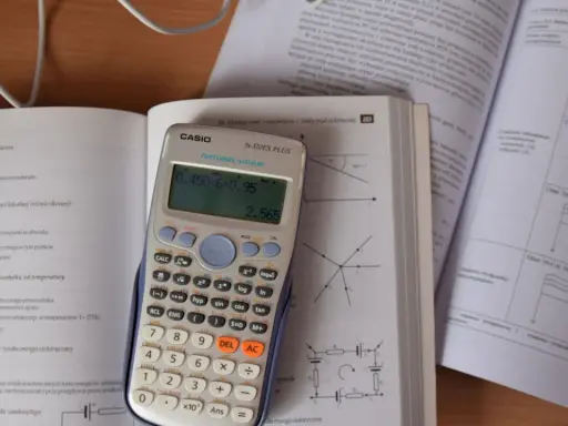Kalkulator Casio z wyświetlonym wynikiem 2.565, obok otwarta książka z fizyki, gdzie można znaleźć informacje o tym, ile to jest kilowat i jak przeliczyć jednostki.