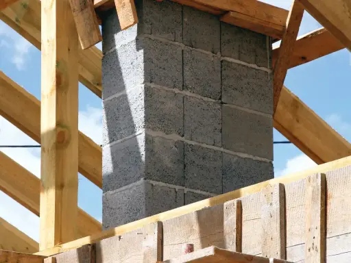 Budowa komina systemowego z bloczków betonowych, gotowego do podłączenia pieca. Drewniana konstrukcja dachu w tle.