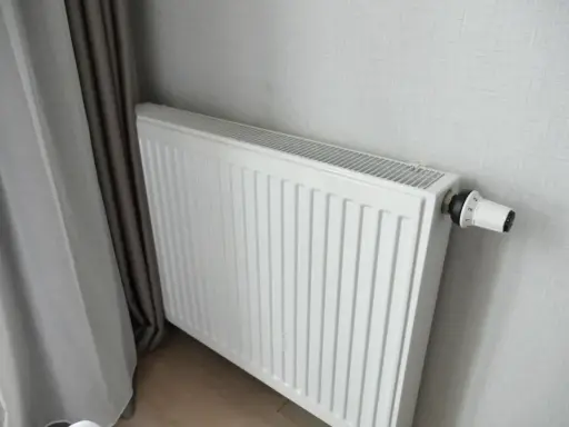Biały grzejnik panelowy z termostatem, idealny do pokoju 16m2. Obok zasłony.