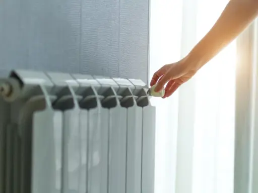 Dłoń reguluje termostat na grzejniku, pokazując różne rodzaje kaloryferów i kontrolę temperatury w domu.