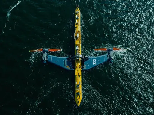 Podwodna turbina wykorzystująca energię pływów, z charakterystycznymi żółto-niebieskimi elementami i pomarańczowymi śmigłami, zanurzona w ciemnych wodach oceanu.
