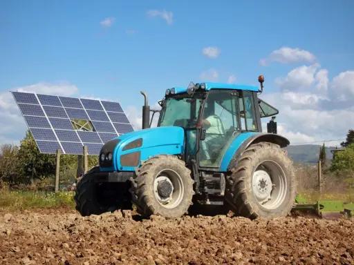 Błękitny traktor pracuje na polu, obok paneli fotowoltaika arimr. Słońce i chmury na niebie.