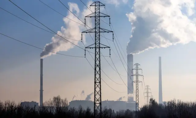 Linie energetyczne i dymiące kominy elektrowni na tle błękitnego nieba. W tle widać kilka wież transmisyjnych i budynki przemysłowe.