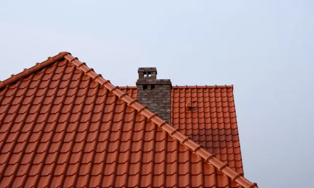 Ceglany komin wystaje ponad dach z czerwonej dachówki. Wlot do komina z pieca znajduje się na wysokości dachu.