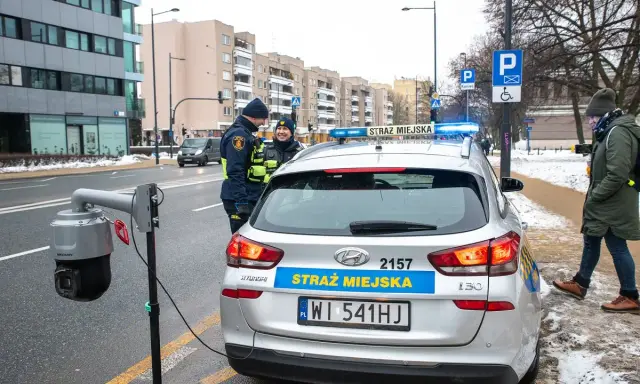 Strażnicy miejscy w Warszawie przy radiowozie i kamerze monitoringu. Dbają o czystą strefę i porządek.